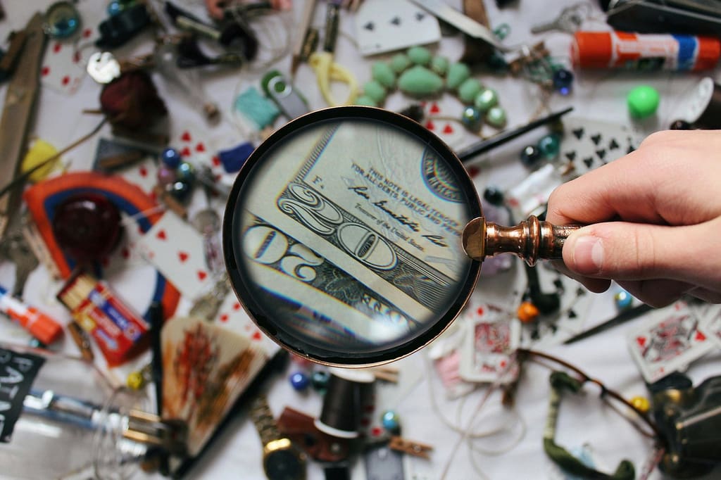 Blog A hand holding a magnifying glass focusing on a twenty dollar bill amidst various scattered objects.